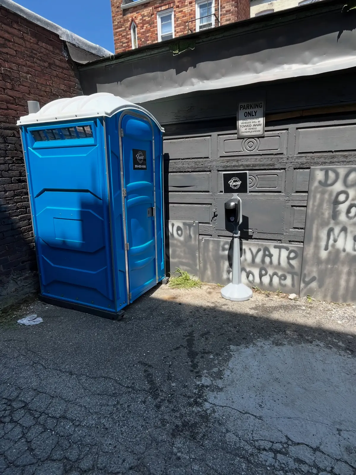 Standard porta potty placed beside a garage on a paved driveway
